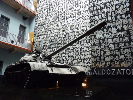 A Soviet tank sits menacingly in a dark space. Behind it, the photographs of the those detained, tortured and killed during oppressive times.