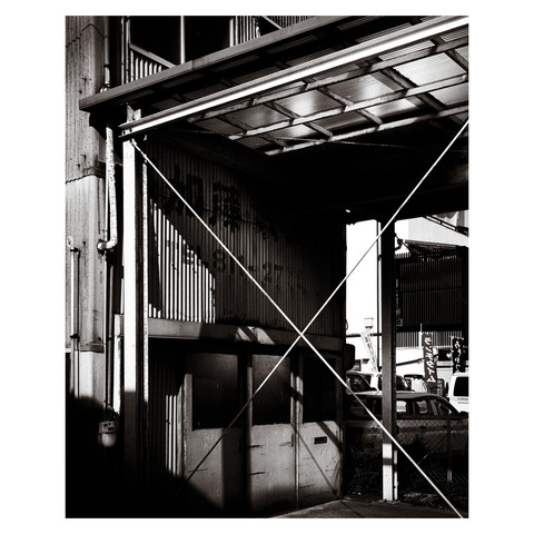 This is a black and white photograph capturing an industrial scene with strong interplays of light and shadow in Nagoya. The focal point is a structure with a corrugated clear acrylic roof that allows the sunlight to filter through, creating a pattern of light and dark on the surfaces below. The textures of the weathered metal walls and the geometric lines of the supporting structures add depth and contrast to the composition. Surrounding elements include glimpses of vehicles and fencing, contributing to the industrial feel of the scene. The photograph captures the essence of a moment of stillness in an otherwise active environment. The monochrome tone conveys a timeless quality, focusing on the interplay of light, materials, and form.
