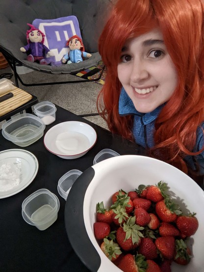 Photo of Fierce wearing a long red wig and a blue sweater, as they're cosplaying Madeline from the videogame Celeste. Fierce is next to a big bowl of strawberries and other cooking utensils. In the background, you can see the plushies of Madeline, Badeline, and the Twitch logo.