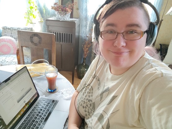 Selfie of a white woman sat at an oval dining table, with a laptop in front of her and a big glass of rosehip soup beside it. There's heavy oak furniture in the background along with frilly cushions, lace curtains, and plants. The room has old lady vibes, because it's the parlour of an OAP obsessed with old fashioned interior design. Jenny, the woman in the photo, smiles a little sleepily at the camera. She's got browny-purply hair, glasses, headphones, and a t-shirt with some sort of big jungle cat on it (badly faded).