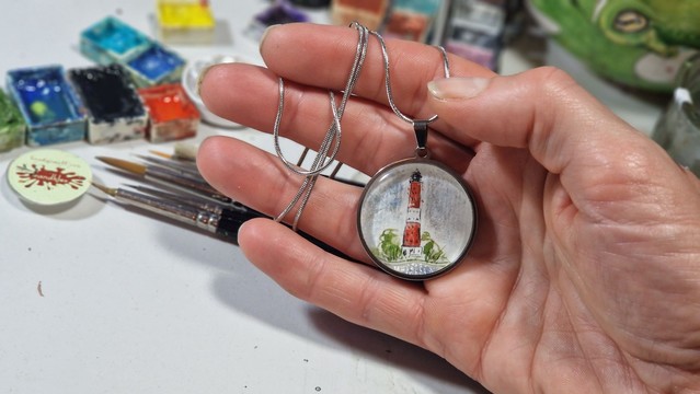 In Silber Anhänger gefasste handgemalte Miniatur des Leuchtturms Pellworm, im Hintergrund Malsachen