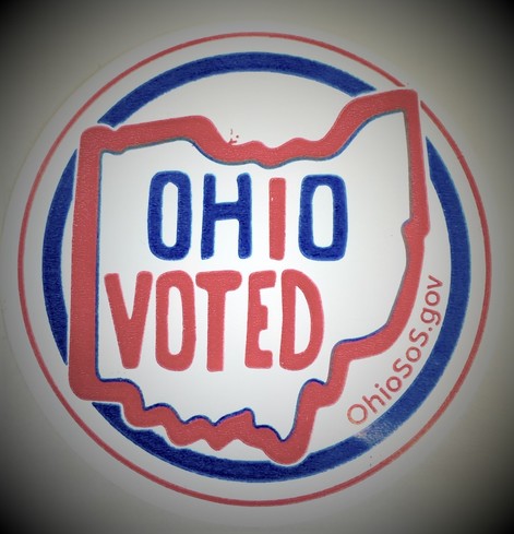 Color image of an "Ohio Voted" sticker, given to Ohio voters at poll locations as they finish their ballots and have them counted. Image is the shape of Ohio in red, outlined by a blue circle, with "Ohio" in blue but for the red "I", and with "VOTED" in red, so it reads both "OHIO VOTED" and "I VOTED OHIO"