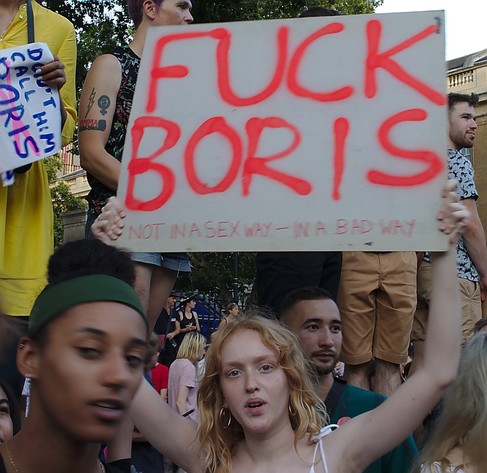 A young woman at a protest holds up a sign that says "Fuck Boris. Not in a sex way - in a bad way"