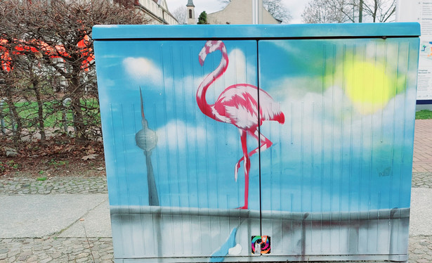 Stromkasten auf einem Bürgersteig mit bisschen kahler Hecke dahinter. Auf dem Kasten ein Bild gesprüht von einem Stück der Berliner Mauer, darüber blauer Himmel, Sonne, weiße Wolken und der Fernsehturm. Auf der Mauer steht ein Flamingo auf einem Bein.
