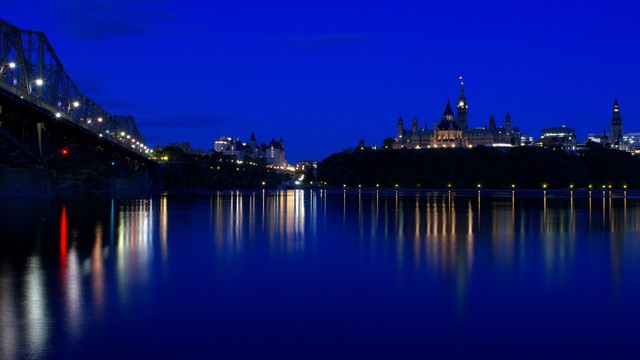 A view of Ottawa's downtown core, as seen from the Quebec side of the Ottawa River, at blue hour.

To the right of the photo, Canada's Parliament Hill is reflected on the water, as are the Rideau Canal and Chateau Laurier, in the centre of the image, and the Alexandra Bridge to the left. Lights leave long streaks in the reflection.

Everything is coated in a deep blue tint.