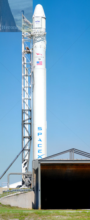 An outdoor, daytime photograph of a tall, white rocket with SPACEX written vertically in blue letters.
On the bottom right corner of the frame is the exhaust vent for the flame trench.