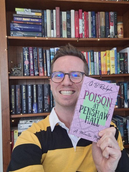 Author G B Ralph holding up a new paperback copy of Poison at Penshaw Hall (A Milverton Mystery) in front of a bookshelf.