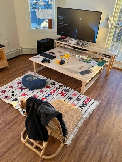 photo of a room with a large monitor with Like A dragon Gaiden video game’s start menu on screen. the monitor is on an ash wood shelf that sits above a shower table made of the same wood. the lower table has peripherals like keyboard, game pad, art supplies, cutting mat, notebooks, mug, etc. A carpet underneath the table has red/black/blue accents, a crescent floor seating pillow, and rattan floor seating beach chair