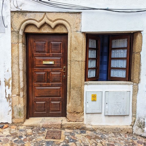 Simple wooden door in an ornate stone frame. There is a window covered in lace curtains to the right of the door.