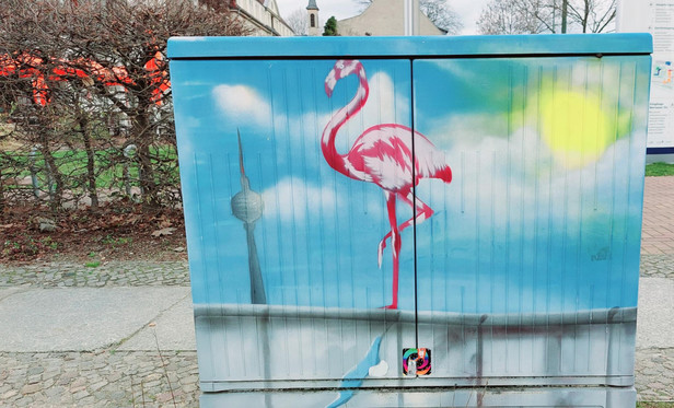 Stromkasten auf einem BÃ¼rgersteig mit bisschen kahler Hecke dahinter. Auf dem Kasten ein Bild gesprÃ¼ht von einem StÃ¼ck der Berliner Mauer, darÃ¼ber blauer Himmel, Sonne, weiÃŸe Wolken und der Fernsehturm. Auf der Mauer steht ein Flamingo auf einem Bein.