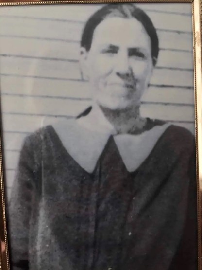 A dark-haired woman in a boxy shirt/dress with a large collar stands in front of a clapboard house. She has a tight lipped smile.