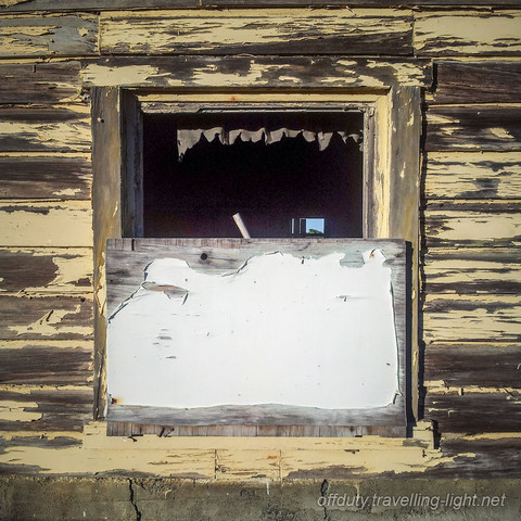 Partly boarded up unglazed window in weatherboard shack, with peeling paint.