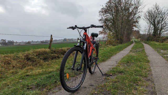 Front view of a red Raleigh Dundee Mountain-Bike on a small famers lane. The sky is grey. 
To the left side is grassland and a wired fence. 
In the background is a hedgerow left and right to the farmers lane.