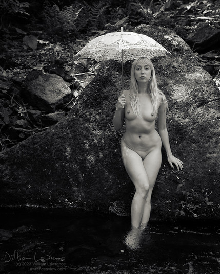 Model Lucy Magdalene stands nude in a stream, leaning against a large rock.  She's holding a parasol and engaging the viewer.