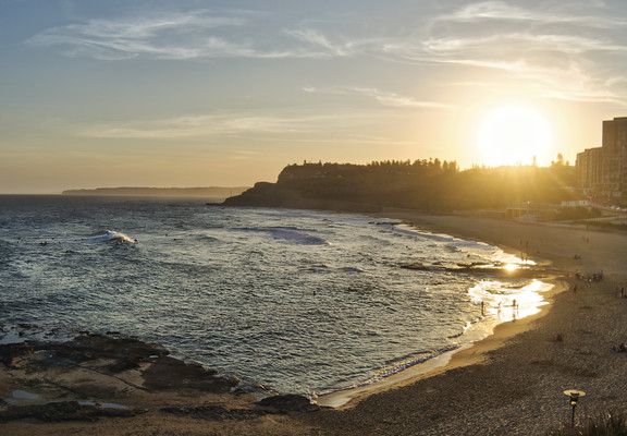 Sunset over a sandy beach cove , with people surfing on the waves, and  playing on the beach