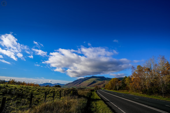 Shot focused on mountain in distance with cloud formation perched above it in blue sky. Foreground of shot is road on right frame and verge/fence on left frame leading towards the mountain