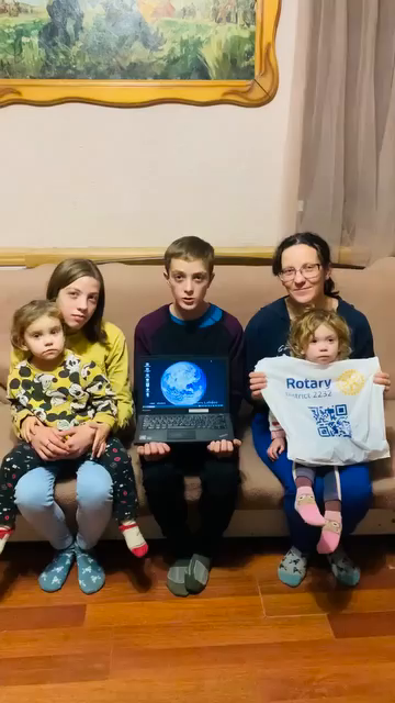 Eine Familie mit 5 Personen sitzen auf einem Sofa, an der Wand dahinter ein Bild. Der etwa 13 Jahre alte Junge hält einen Laptop aufgeklappt auf den Knien. Rechts und links davon die Mutter und Geschwister. Ein Kind hält noch eine Tüte mit Rotary-Logo in die Kamera.