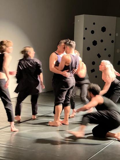 Two figures hold each other center. Five blurred bodies circle the pair. In a white theatre with gray floors, bright light. A wall at rear has numerous mirrors in varying shapes and sizes.