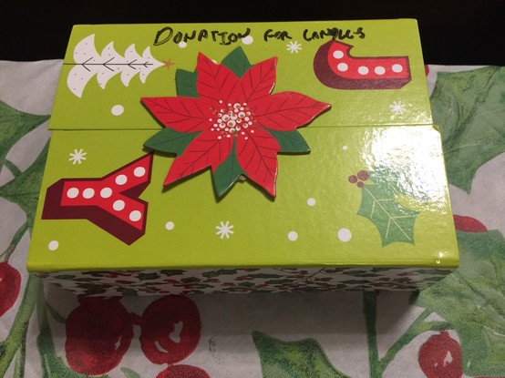 Color image of a decorated donation box at an inner-city church in Northeast Ohio, USA, to fund advent candles. The box is a festive light green, with depictions of trees and holly, with the letters JOY spelled out diagonally on the top, the O depicted by a poinsettia bloom  with read and green leaves.