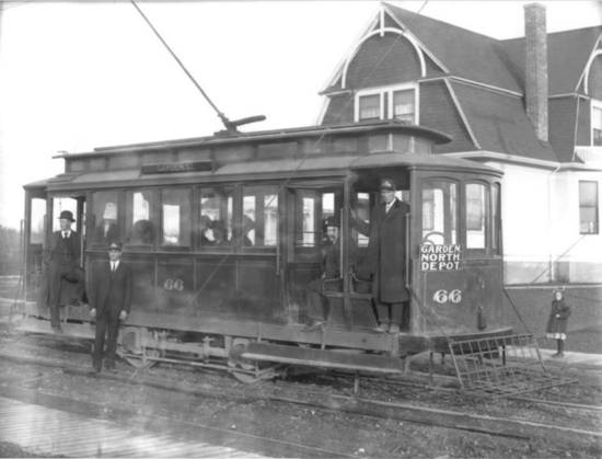 Black and white photo circa 1906-1909 of an electric trolley in front of a residential house. Sign on the front of the trolley reads "Garden North Depot - 66" One uniformed trolley employee stands at the front entrance of the trolley and another stands on the tracks beside it. A well-dressed man in a suit, bowler hat, and overcoat stands on the rear running board, hand in pocket, staring into the distance.