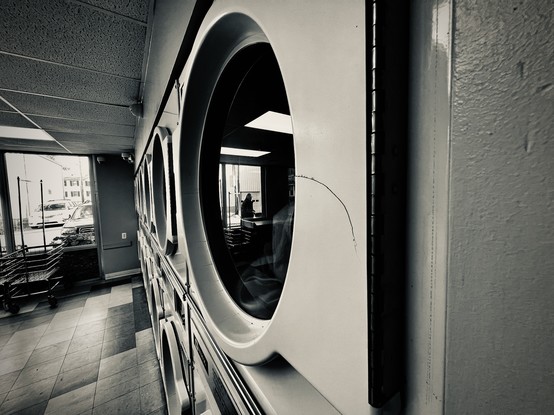 A row of washers and dryers in a laundromat with multicolor tile floors, drop ceilings, and a crack in the plastic facade surrounding the dryer door. Shot at a shallow angle, catching the reflection of someone standing outside talking on their phone.