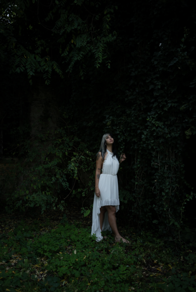a girl standing near a ivy covered wall, with a white long dress, watching above her head, the left arm slightly raised