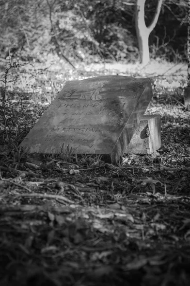 A fallen tombstone in the shape of a cross lies on another tombstone on an abandoned cemetery.