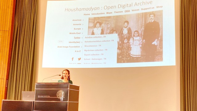 Dr. Lévy-Aksu stands at a podium in front of a slide of her presentation. The slide reads" "Houshamadyan: Open Digital Archive"