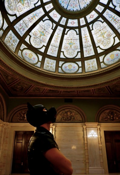 A hooded human dog in leather crosses his arms underneath a glorious dome of Tiffany glass.
