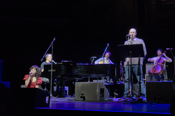 Ben Folds playing piano on stage at the Royal Albert Hall, along with Matt Lucas stood singing, a woman crouched singing, and a cellist and guitarist playing in the background.