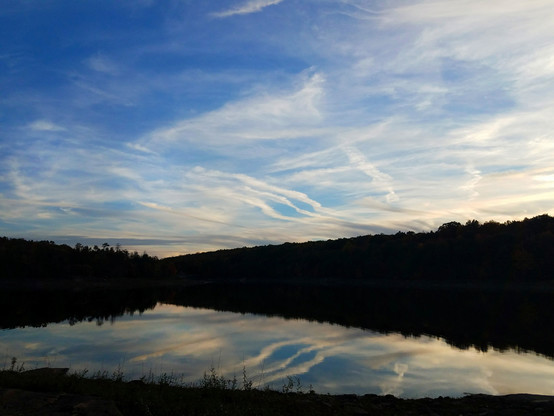 On the shore of a lake. Low wooded ridges rise above the far shore. All land features are silhouetted in the twilight. The sky is a deep blue with streaks of white cloud mingling with airplane contrails from a nearby small airport, lingering and slowly spreading until they become almost indistinguishable from the streaky clouds.