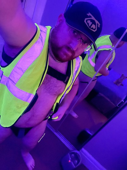 A selfie from arms length looking into the camera, wearing a nasty pig hat, my neoprene harness, hi-viz vest, and leather jockstrap. My stuffed little coyote sits on the bed reflecting in the closet mirror behind me.