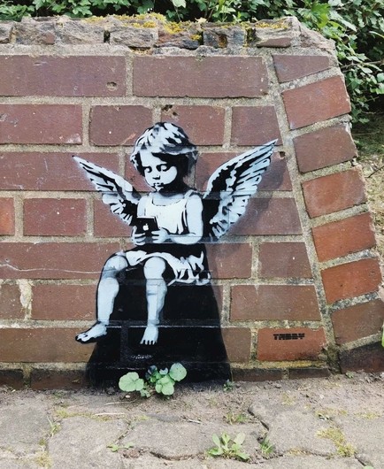 Streetartwall. The black and white mural of a little angel has been sprayed/painted on a low red brick wall. He has light-colored curly hair, wears a simple white shirt, wings and is barefoot. He sits on a black stone and looks with interest at an Apple iPhone.