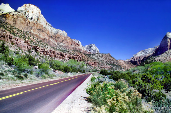 Beneath a deep blue cloudless desert sky, a scenic 2-lane highway following a semiarid river valley through rugged mountains. It is spring and recent rains have brought out vivid green leaves on some of the trees and shrubs. Shrubs with pale yellow blossoms line the roadside on our right. To our left a mostly barren steep mountainside rises sharply not far from the roadside. It is sprinkled with scattered olive green scrubby vegetation in a wide swath between stark naked rock cliffs above and below.