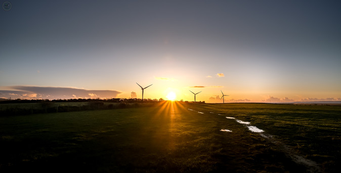 Sunset behind wind turbines, fields and hedges in foreground, clear skies with scattered clouds, sunrays in centre of shot