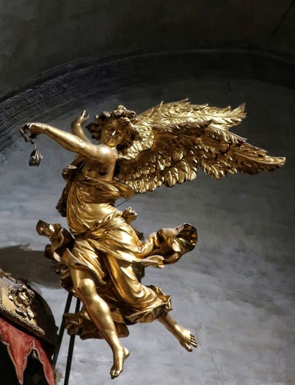 A dancing golden baroque angel against a dark grey wall.