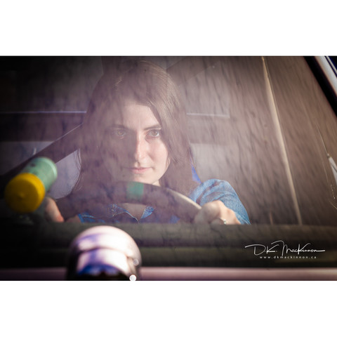 dark-haired woman on a denim dress behind the wheel of a vintage stock car, looking through the windshield.
