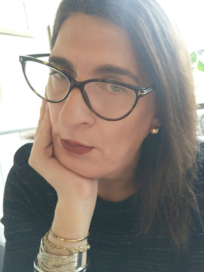 A selfie of a brunette trans women wearing glasses, gazing into the camera while supporting her chin on her hand, the arm festoomed with bangles