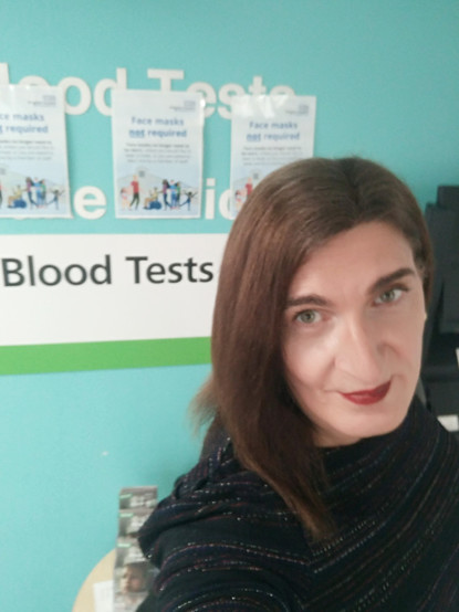 A selfie of a brunette trans woman standing next to a sign saying "Blood Tests"
