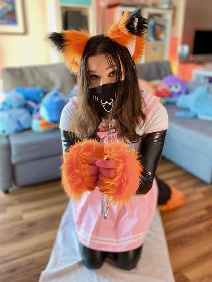 Rina kneeling on floor, offering her collar and leash to the camera. She is wearing a pink and white Japanese schoolgirl uniform over a black latex catsuit, with orange and black fox ears, paws, and tail