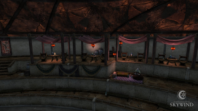 Canopied seating areas are displayed. There are two chairs and a table with drinks under each canopy, which are red in color. There are also hanging red lanterns under each canopy. On the tier below the canopy level, a Dunmer sits cross legged on a rug surrounded by pillows.