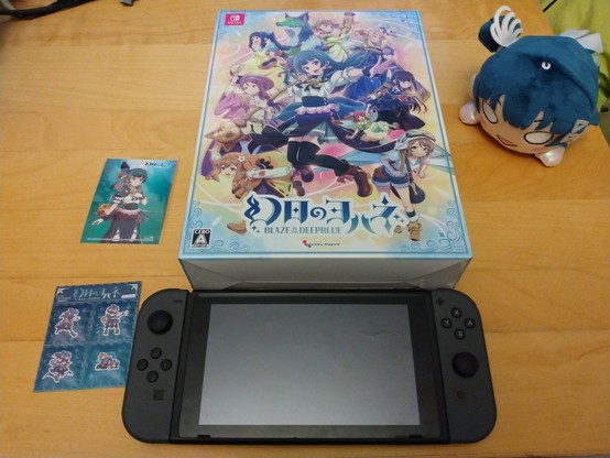Box containing a Switch copy of Genjitsu no Yohane: Blaze in the Deepblue. Below it is a black Switch. Beside it are a picture of Yohane, fridge magnets with pictures of Yohane, and a Yohane nesoberi.