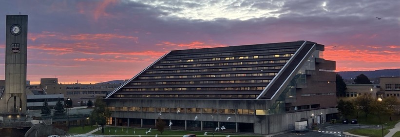 A colourful sunrise sky over the main library at Memorial University (St. John's (NL) Canada. The library building has rows of lit windows that follow a slanted, stair-step look. There is a lit clock tower to the left, with sidewalks and a sparsely filled parking lot in front. The sky has brilliant reds near the horizon, with pink highlights on the underside of dark purple clouds. A small patch of light blue sky hovers at the top centre of the photo. (November 2023]