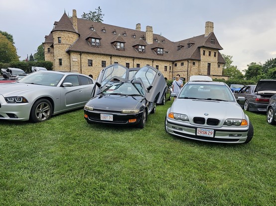 Black small car with open dihedral doors ("butterfly doors") on the grass amongst many other vehicles, in front of a castle-like mansion (the Larz Anderson Auto Museum in Brookline, Massachusetts).