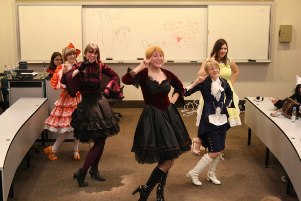 Five dancers (in mostly Japanese loli style costumes of a rainbow color) pose after a performance in a university classroom during an anime convention.