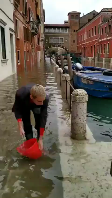Man standing on a flooded sidewalk using a bucket to futilely bail water into the canal that is flooding the sidewalk.