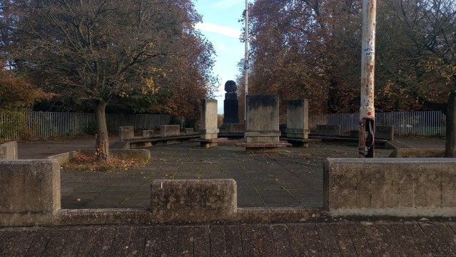 A broad avenue, perhaps, interrupted by a circle of low grey concrete barriers topped with irregular castellations that might, or might not, serve as backrests should the barriers be used for seating.

In the centre of the circle are three rectangular monoliths, roughly four times the size of tombstones, which each are skirted by flat wooden benches, presumably in the hope that people might gather here in happy, chattering crowds.

There are no happy, chattering crowds. Just a lamp-post, rusted and stickered, which has lost an inspection cover so that its meagre electrical entrails are exposed, and a small and straggly tree, looking as resignedly neglected as the whole.

On the far side of the circle, silhouetted against a washed-out sliver of blue sky, is the back of a vast stone head mounted on a  massive rectangular plinth.