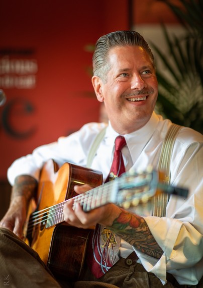 A 1920s style dressed man (Paul) with a 1920s style haircut and mustache, with a Selmer guitar and a big smile on his face.