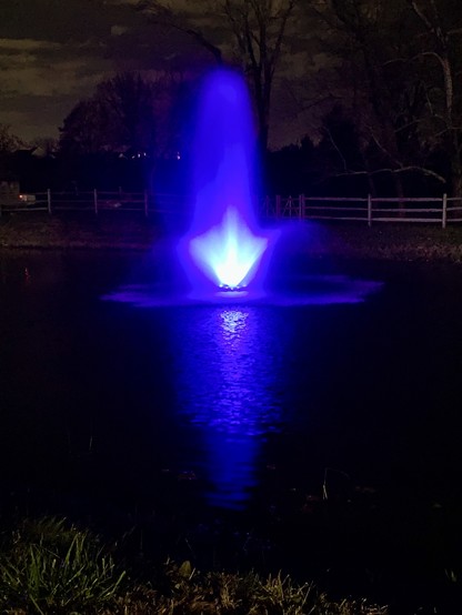 A water fountain in a pond is lit up by a blue light at night.