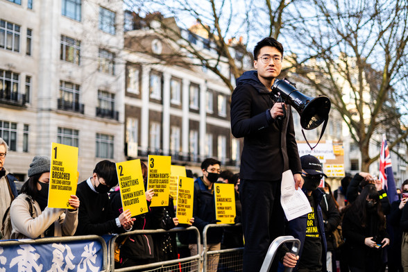 Portrait of Simon Cheng speaking at a rally outside the Chinese Embassy in London. Cheng is holding a megaphone atop a small step ladder. In the background are other supporters of the Hong Kong pro-democracy movement holding placards showing the "five demands" in black text on a yellow background.